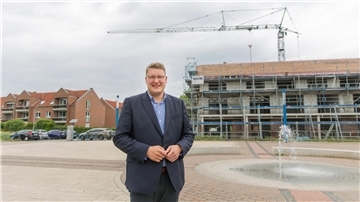 Ein Mann steht auf einem gepflasterten Platz. Im Hintergrund ein Gebäude im Bau mit Kran.
