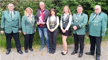 Gruppenfoto von sieben Mitgliedern eines Schützenvereins in Uniform oder mit Schützenorden, aufgenommen im Freien vor grüner Hecke. Eine Person hält ein Geschenk.