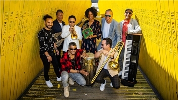 Die achtköpfige Band „CaboCubaJazz“ posiert fröhlich in einem gelben Durchgang. Die Musiker halten Instrumente wie Trompete, Conga, Posaune und Keyboard.