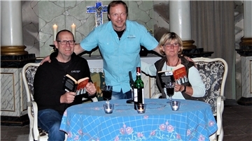 Eine Frau und ein Mann sitzen an einem Tisch. Sie halten jeweils ein Buch in der Hand. Ein weiterer Mann steht zwischen den beiden.