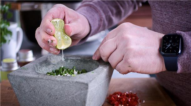 Christian Schwarz presst eine ganze Limette über die Zutaten und zerreibt sie mit dem Mörser zu einem Brei. Dann folgen der Knoblauch und ein Schuss Olivenöl. Nach einer weiteren Runde im Mörser gibt der Grill-Influencer je einen Teelöffel rosa Himalaja-Salzflocken, Pfeffer, Oregano und Thymian hinzu. Noch einmal wird alles gemörsert, dann kommt die Chimichurri ins Einmachglas. Er füllt das Glas mit Olivenöl auf, bis die Paste gerade so bedeckt ist. Jetzt fehlt nur noch das Lorbeerblatt, dann ist die Steak-Sauce fertig.