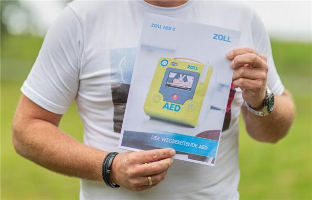 Ein Zettel, auf dem ein Defibrillator abgebildet ist, wird von einem Mann in die Kamera gehalten. Zu sehen sind nur die Arme und der Bau des Mannes, alles andere ist "abgeschnitten".