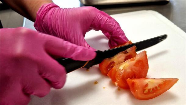 Ein Mensch schneidet mit einem Messer eine Tomate - dabei trägt er Einweg-Handschuhe.
