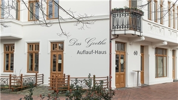 Eine weiße Gründerzeitfassade mit Holzfenstern und Türen und der Aufschrift "Das Goethe Auflauf-Haus".
