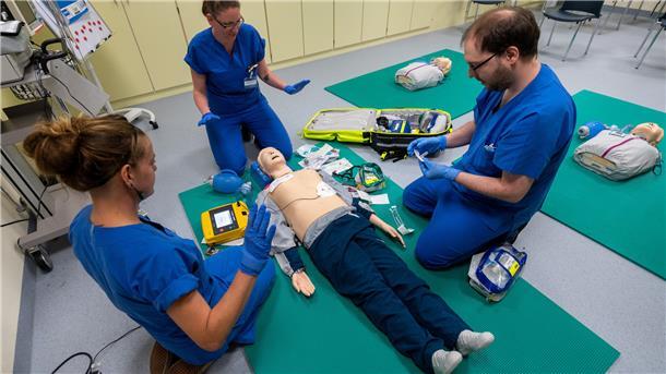 Drei angehende Ärzte und eine Puppe, die auf dem Boden liegt, sind zu sehen. Es geht um das Thema Humanmedizin.