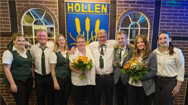 **ALT-Text:**
Gruppenfoto des neuen Königshauses des Schützenvereins Hollen: Acht Personen, teils in Uniform, stehen vor einem Schild mit der Aufschrift „Hollen“, zwei von ihnen halten Blumensträuße.
