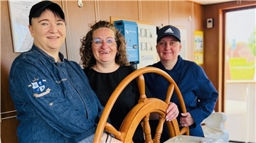 Drei Frauen stehen lächelnd an einem hölzernen Steuerrad auf der "MS Hansa"