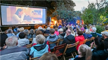 Menschen sitzen auf Stühlen im Freien vor einer Leinwand.