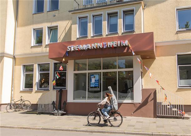 Ein gelbes Gebäude, an dem eine Frau auf einem Fahrrad vorbeifährt. Über dem Eingang steht "Seemannsheim".