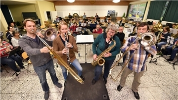 Musikerinnen und Musiker des Sinfonischen Blasorchesters Wehdel proben gemeinsam in einem Saal. Im Vordergrund spielen zwei Posaunisten, eine Klarinettistin und eine Saxofonistin, während im Hintergrund das gesamte Orchester mit Blech‑, Holzbläsern und Schlagwerk zu sehen ist.