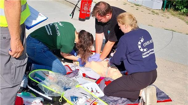 Drei Mitglieder des Jugendrotkreuzes üben an einer Übungspuppe eine Reanimation, während ein Schiedsrichter mit Klemmbrett danebensteht.