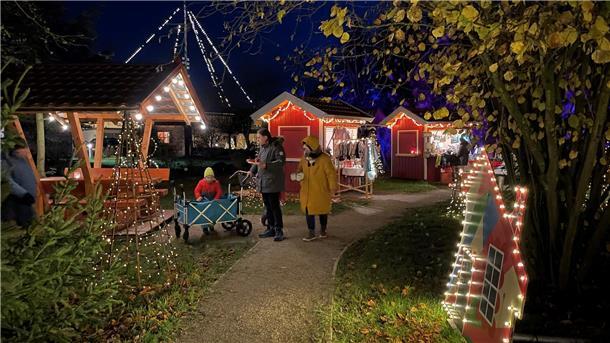 Mit Lichterketten geschmückte Holzhütten in einer Gartenanlage; dazwischen Menschen auf Kiesweg.