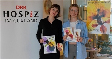 Zwei junge Frauen stehen nebeneinander vor einem Roll-up des DRK-Hospiz im Cuxland und halten Materialien der Kampagne „Zwischen Anfang und Ende“ in den Händen. Im Hintergrund ist ein farbiges Kampagnenplakat mit Blütenmotiv zu sehen.