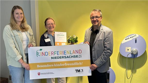 Drei Personen halten eine Urkunde und ein Banner mit der Aufschrift „Kinderferienland Niedersachsen – Besonders kinderfreundlich!“ vor einer gelben Wand.