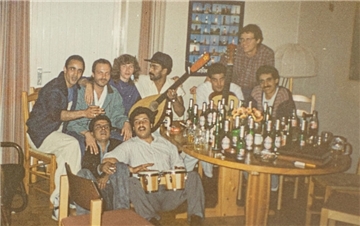 Eine Gruppe Männer mit einer Frau in ihren Reihen. Sie sitzen an einem Tisch, der vollgestellt ist mit Bierflaschen. Ein Mann hält eine Oud in den Händen, ein anderer eine Gitarre, ein dritter zwei Handtrommeln (vielleicht eine Darbuka).