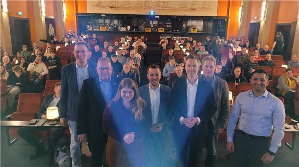Gruppenfoto bei einer Podiumsdiskussion in einem historischen Kinosaal mit maritimer Wandmalerei. Im Vordergrund stehen sieben Personen, darunter Politiker und Veranstalter, vor einem Publikum aus Schülern und Erwachsenen, das in den roten Kinosesseln sitzt.