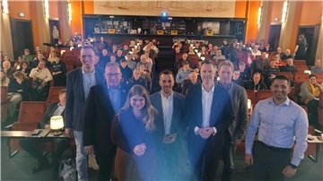 Gruppenfoto bei einer Podiumsdiskussion in einem historischen Kinosaal mit maritimer Wandmalerei. Im Vordergrund stehen sieben Personen, darunter Politiker und Veranstalter, vor einem Publikum aus Schülern und Erwachsenen, das in den roten Kinosesseln sitzt.