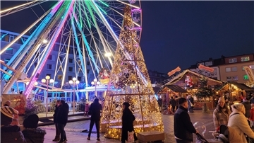 Riesenrad und Lichtkegel zwischen Buden auf dem Weihnachtsmarkt; dazwischen Menschen.