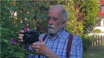 Ein älterer Mann mit Brille und grauem Bart fotografiert mit einer Kamera Pflanzen im Garten.