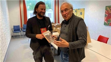 Zwei Männer stehen in einem hellen Raum mit Tischen und Stühlen. Beide Männer halten Bücher in den Händen und lächeln. Im Hintergrund sind bunte Bilder an der Wand und gepackte Kartons auf einem Tisch zu sehen.