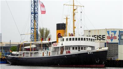 Viel Dampf, wenig Geld: Eisbrecher „Wal“ braucht weiter finanzielle Hilfe Der historische Dampfeisbrecher „Wal“ liegt im Hafen vor einer Werftanlage. Das Schiff ist schwarz-weiß mit einem markanten gelb-schwarzen Schornstein. Rettungsringe hängen an der Reling, und im Hintergrund sind Kräne sowie industrielle Gebäude mit Werbeaufschriften zu sehen.
