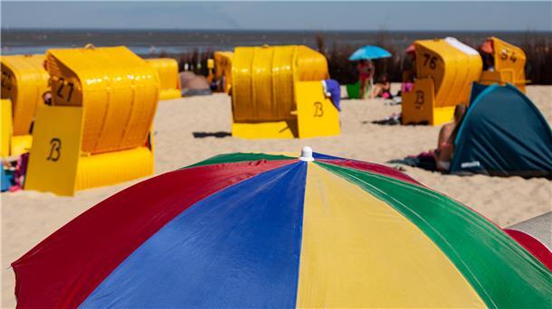 Ein bunter Sonnenschirm im Vordergrund, dahinter Sandstrand, gelbe Strandkörbe und Menschen, verschwommen.