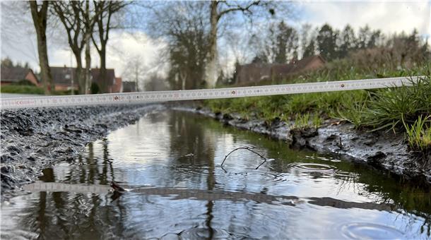 Aufgefahrener Seitenraum einer Landesstraße, in dem sich Wasser sammelt Von links nach rechts liegt ein Zollstock. Der Seitenraum liegt höher als die Straße.