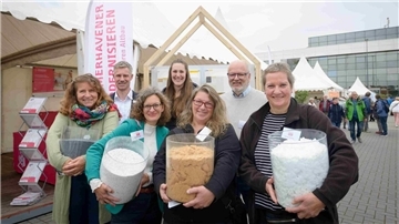 Gruppe von sieben lächelnden Personen beim Energie- und Klimastadttag in Bremerhaven, die verschiedene Dämmmaterialien in transparenten Behältern präsentieren. Im Hintergrund sind Infostände, ein Holzrahmenmodell und Besucher des Aktionstags zu sehen.