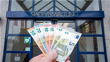 Eine Hand hält Geldscheine vor einer Tür, über der Stadtverwaltung geschrieben steht.