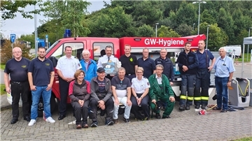 Menschen sitzen vor einem Feuerwehr-Fahrzeug auf dem "GW-Hygiene" steht. 