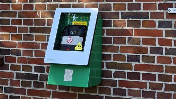 Hinter einer Scheibe in einem grünen Kasten ist ein Defibrillator sichtbar.