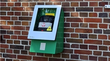 Hinter einer Scheibe in einem grünen Kasten ist ein Defibrillator sichtbar.