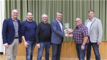 Eine Gruppe von sechs Männern steht nebeneinander in einem Raum mit Holzboden und grünem Vorhang im Hintergrund. Zwei Männer in der Mitte schütteln sich die Hand; einer von ihnen überreicht ein Geschenk.
