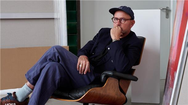 Ein Mann mit Brille, dunkler Jacke und Mütze sitzt nachdenklich in einem Designerstuhl. Er hat ein Bein über das andere geschlagen und stützt sein Kinn auf die Hand. Im Hintergrund sind Leinwände und ein Kunstwerk zu sehen.