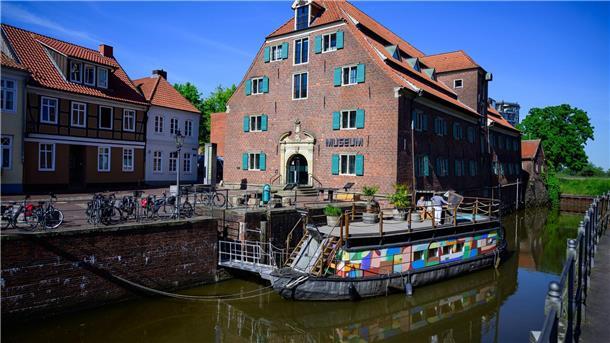 Ein kleines Schiff mit bunten Seitenwänden liegt in einem Hafen. Im Hintergrund sind alte Backsteingebäude zu sehen.  