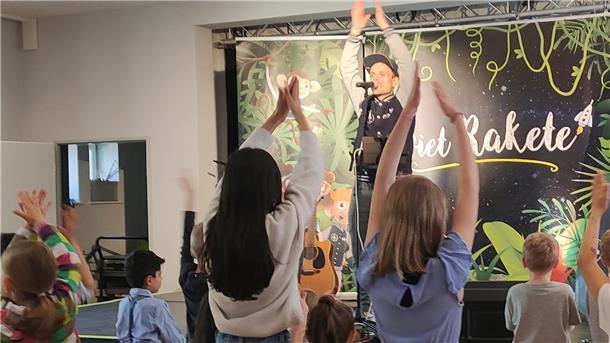 Verschiedene Kulturen treffen beim Frühlingsfest im Jugendhaus aufeinander Ein Mann steht auf einer Bühne und hebt die Hände, mehrere Kinder stehen davor.