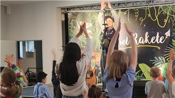 Ein Mann steht auf einer Bühne und hebt die Hände, mehrere Kinder stehen davor.