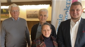 Vier Personen stehen in einem Innenraum vor einer Wand mit beleuchteten Bildern und einer Fahne mit der Aufschrift „Freie Demokraten“. Die Gruppe besteht aus zwei älteren Männern, einer Frau mit Brille und einem jüngeren Mann im Anzug.