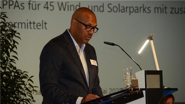 PNE AG trotz Verlusten auf Wachstumskurs mit Wind- und Solarenergie **ALT-Text:**
Ein Redner spricht bei einer Präsentation am Mikrofon, vor sich ein Glas Wasser und ein Rednerpult, im Hintergrund ist ein Text zur Energiewirtschaft projiziert.