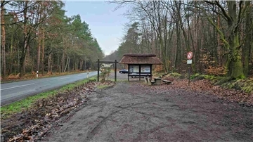 Redakteur sagte:
Parkplatz „Steingarten“ an der K21 im Waldgebiet bei Lamstedt mit Infotafel, Reetdach-Unterstand, Sitzgruppe und angrenzender Landstraße, im Hintergrund ein geparktes Auto.