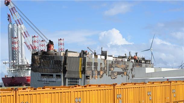 Fremantle Highway: Ladung wird nach Feuer vom Autofrachtschiff geborgen Der schwer beschädigte Autofrachter „Fremantle Highway“ liegt im Hafen, abgeschirmt durch gelbe Container.