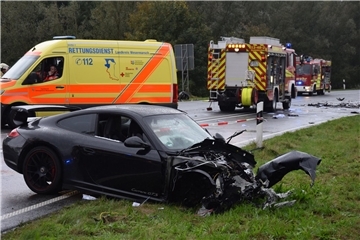Ein Foto von einer Unfallstelle. Im Vordergrund: ein schwarzes Auto, das vorn stark zerstört ist. Im Hintergrund sind ein Krankenwagen und zwei Fahrzeuge der Feuerwehr zu sehen. Der Unfall ereignete sich  7. Oktober 2023 auf der Bundesstraße 212 und forderte zwei Todesopfer. Nun prüfen Sachverständige erneut, ob der Porsche-Fahrer tatsächlich die Ursache war.