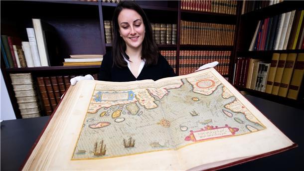 Eine Frau hält mit Handschuhen einen sehr großen und alten Seeatlas in ihren Händen