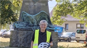 Ein Mann in einer signalgelben Weste und einem Helm in der Hand steht vor einem Denkmal.