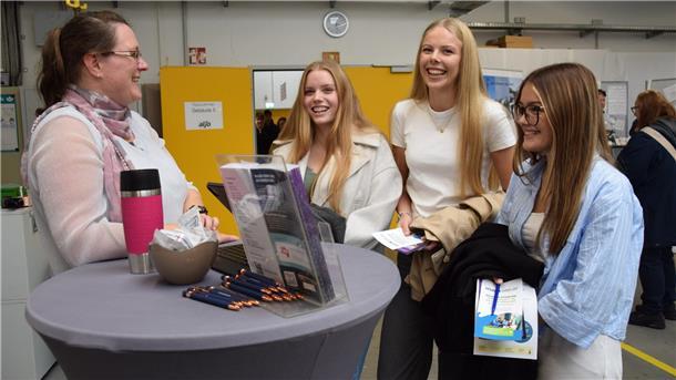 Drei junge Frauen stehen vor einem Info-Stand. Links im Bild steht eine Frau am Tisch, die die Frauen berät. 