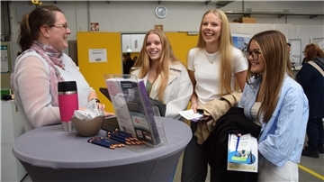 Drei junge Frauen stehen vor einem Info-Stand. Links im Bild steht eine Frau am Tisch, die die Frauen berät. 