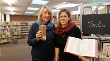 Zwei Frauen stehen in einer Bibliothek, mit jeweils einem Buch in der Hand.