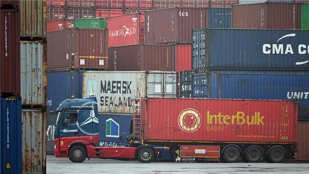 Ein Sattelzug mit einem roten Container der Marke 'InterBulk Group' steht auf einem Hafengelände, umgeben von gestapelten Frachtcontainern in verschiedenen Farben und mit Markenaufdrucken wie 'Maersk Sealand' und 'CMA CGM'.