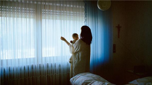 Eine Frau steht an einem Fenster. Im Arm hält sie ein Kind. 