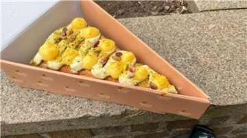 Ein Stück Croissant-Pizza mit Creme und Pistazien in einer rosa Box auf Steinboden.